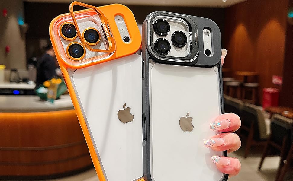 A hand holding two smartphone cases, one orange with a clear back and MagSafe compatibility, and the other black with a clear back. Both cases feature cutouts for dual cameras and an Apple logo in the center. The background shows a café setting with blurred furniture.