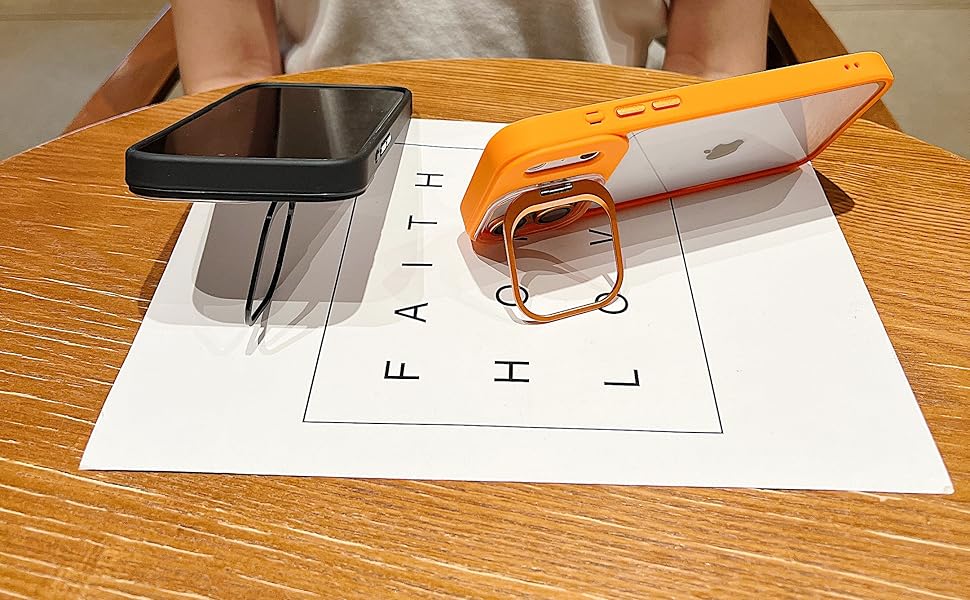 Two smartphone cases resting on a white piece of paper with the word "FAITH" printed on it. One case is black and flat, while the other is orange and clear with a MagSafe design. The wooden table surface and a person sitting in the background are visible.