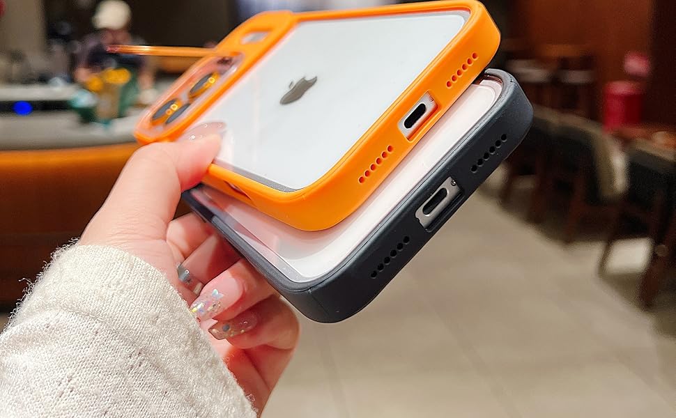 A hand holding two smartphone cases stacked on top of each other. The top case is orange and clear, designed for MagSafe compatibility, while the bottom case is black. The scene is set in a café with blurred background details.